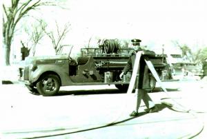 black and white photo of a man holding a hose in front of an old fire truck